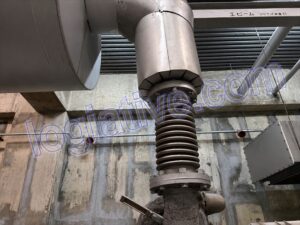 0d1be9a4-245c-43e6-a610-00ebfde6ed39_R / 大型発電機・非常用発電機買取.com Close-up of a heavy-duty screw-actuated actuator with a ribbed threaded column and flanged joint, mounted to a large industrial pipe in a concrete plant setting.