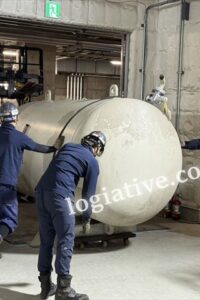 Four workers in blue coveralls and hard hats push a large white spherical tank on a wheeled cradle inside an industrial facility.